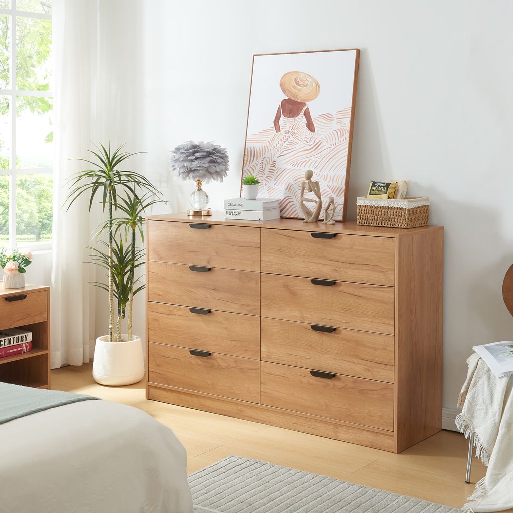 A bedroom scene featuring a wooden dresser with drawers, a lamp, plants, and artwork.