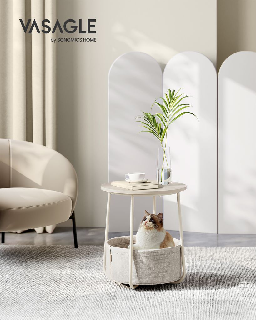A cat sits in a round basket-style side table, with a chair and decorative screens in the background.