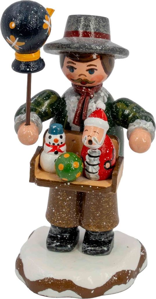 Ein handgefertigter Holzfigur zeigt einen Spielwarenhändler mit einem Tablett voller bunter Spielzeuge in den Händen und einem Vogel auf einem Stock während er auf einem schneebedeckten Sockel steht