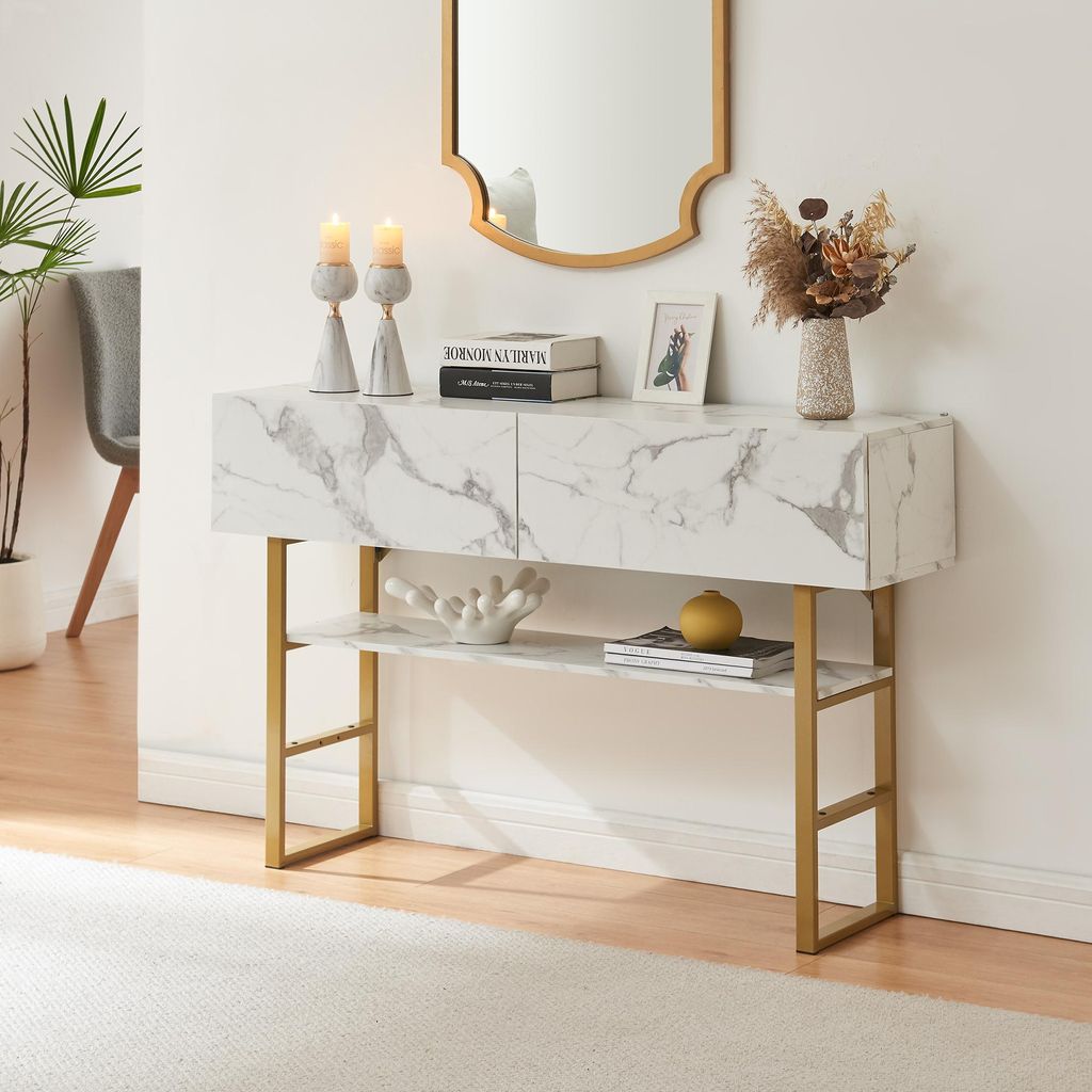A console table with a marble-like finish and gold legs, decorated with candles, books, a vase with dried flowers, and a decorative object.