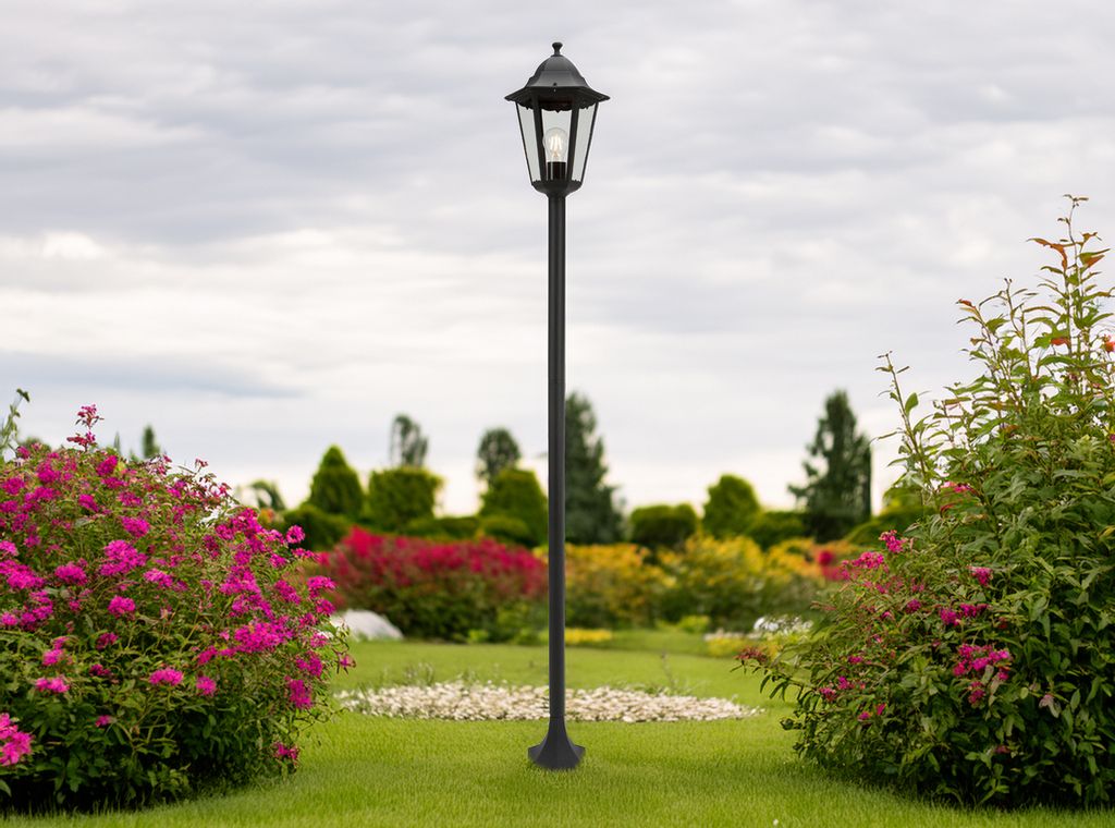 A black garden lantern stands in a lush garden with blooming pink flowers and green bushes.