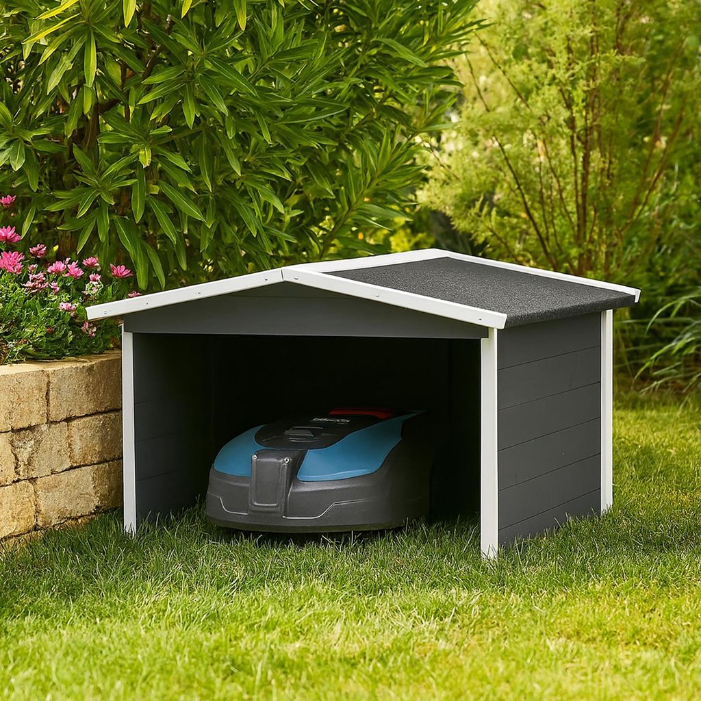 A robotic lawnmower is parked inside a small, dark gray shed with a white trim and a dark gray roof. The shed is situated on a green lawn, with a stone wall and pink flowers to the left, and trees and foliage in the background.