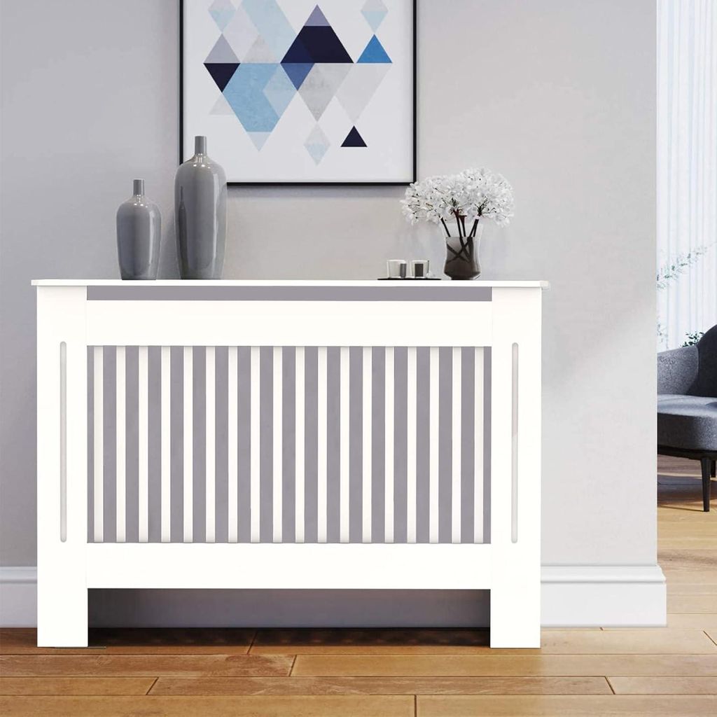 A white radiator cover with vertical gray slats is shown in a room setting, with decorative vases and a framed picture on top.