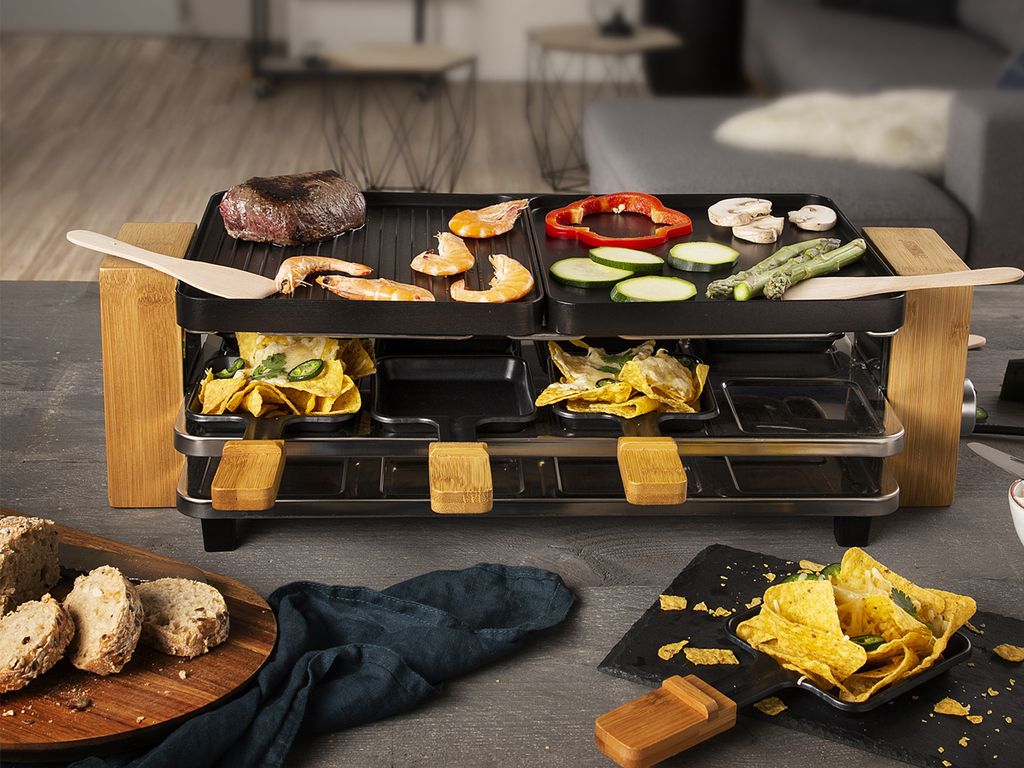 A raclette grill is set up on a table with food being cooked on it, and bread and chips are placed nearby.