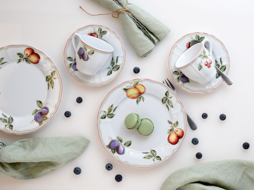 A top-down view of a fruit-themed porcelain tea set with two cups and saucers, three plates, and two macarons, arranged on a white background with scattered blueberries and green napkins.