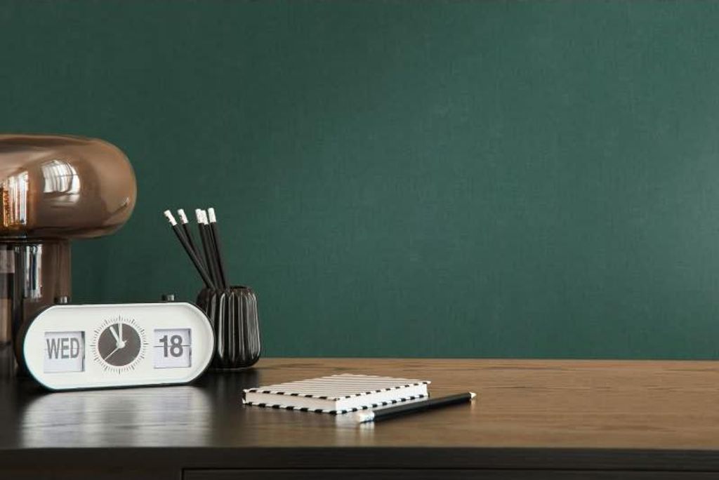 A desk with a clock, notebook, pencils, and a lamp against a dark green wall.