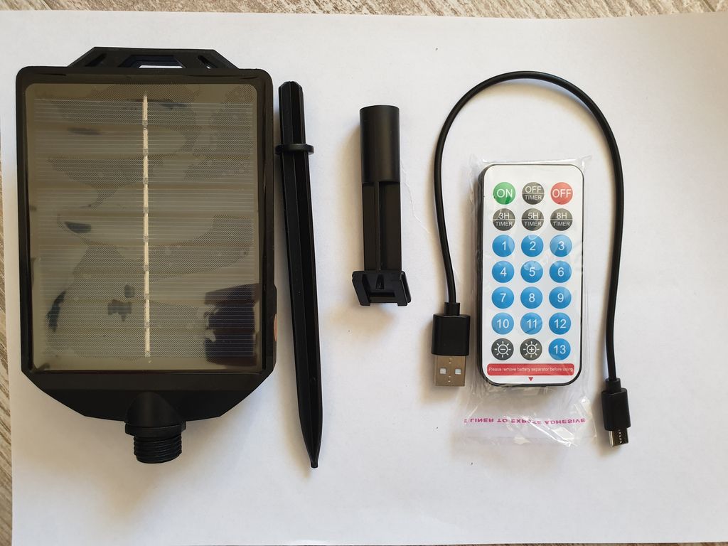 A flat lay of a solar panel, a stake, a remote control, and a USB cable on a textured surface.