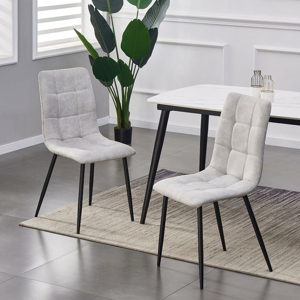 Two dining chairs are shown in a room setting, with a table and plant in the background.