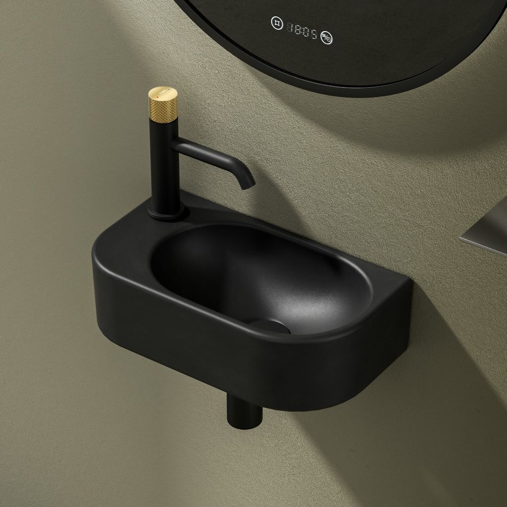 A black wall-mounted sink with a black faucet and a gold accent on the faucet handle, set against a textured greenish-gray wall. A portion of a circular mirror with digital display is visible in the upper background.