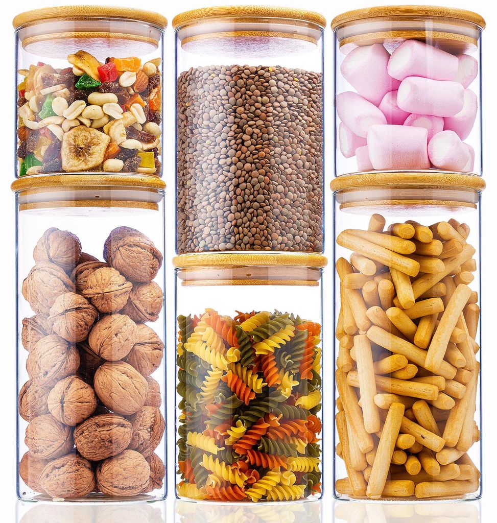 Sechs klare Glasvorratsgläser mit hellen Bambusdeckeln, gefüllt mit verschiedenen Trockenwaren wie Nüssen, Linsen, Marshmallows, Nudeln und Brotstangen