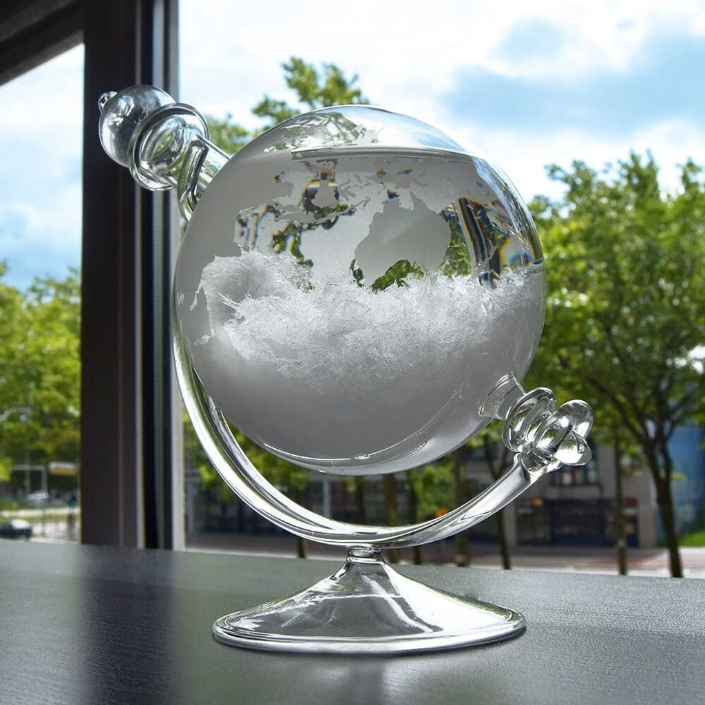 A glass globe-shaped barometer with a map of the world etched on it, filled with white crystals, sits on a dark surface in front of a window with trees and sky visible outside.