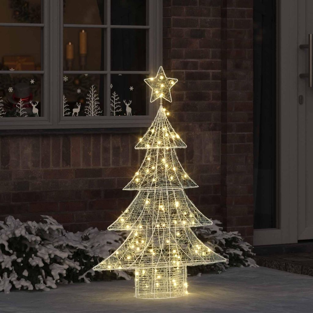 A lighted Christmas tree decoration stands outside a brick building next to a window.