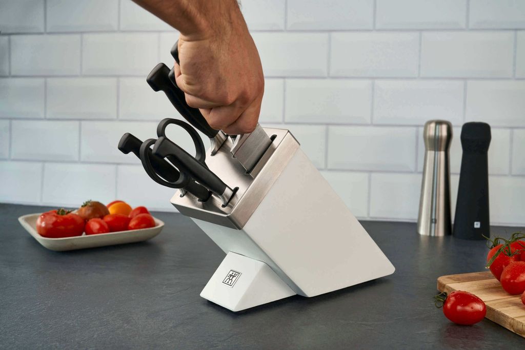 A hand is placing a knife into a white knife block on a kitchen counter, with tomatoes in a dish and salt and pepper grinders in the background.