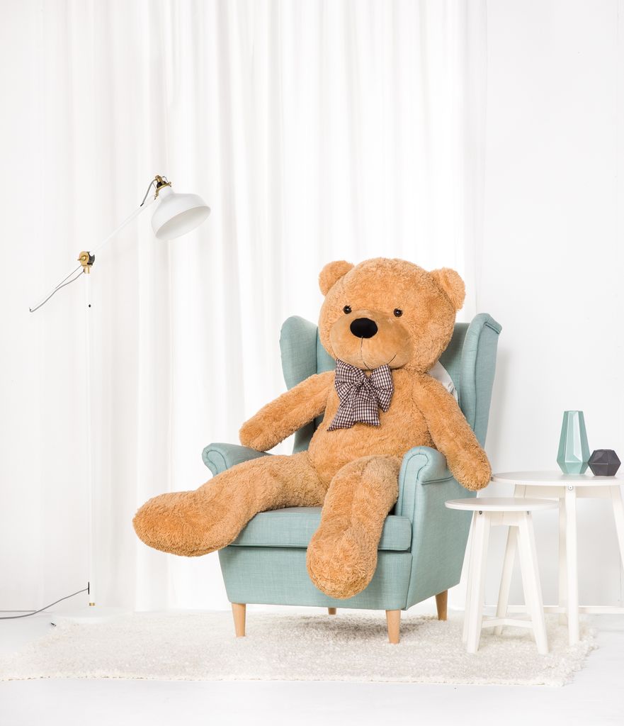 A large teddy bear is sitting in a light blue armchair in a room with white curtains.