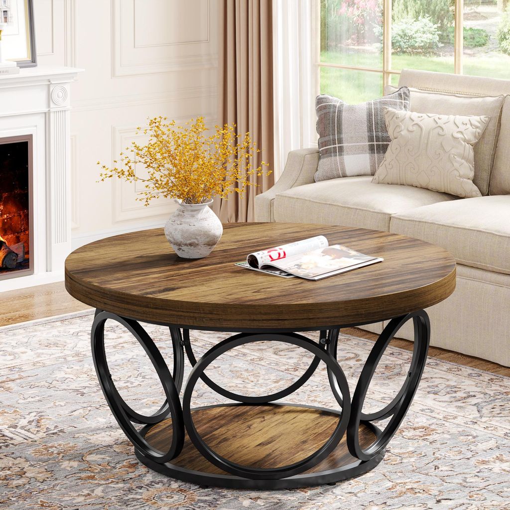 A round coffee table with a wooden top and a black metal base with circular accents, placed in a living room setting with a fireplace and sofa in the background.