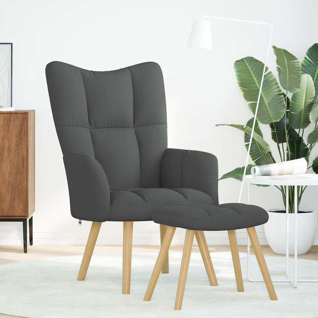 A dark gray fabric armchair with a matching ottoman, placed on a light-colored rug in a well-lit room with a large plant and a white side table.