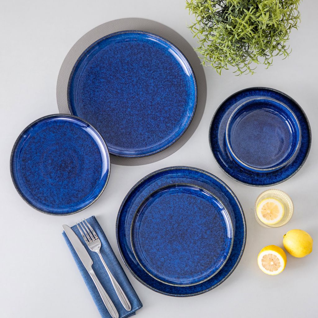 A collection of blue ceramic plates and bowls arranged on a light gray surface, with two lemon halves and a small glass of lemon water in the foreground, and a green plant in the background.