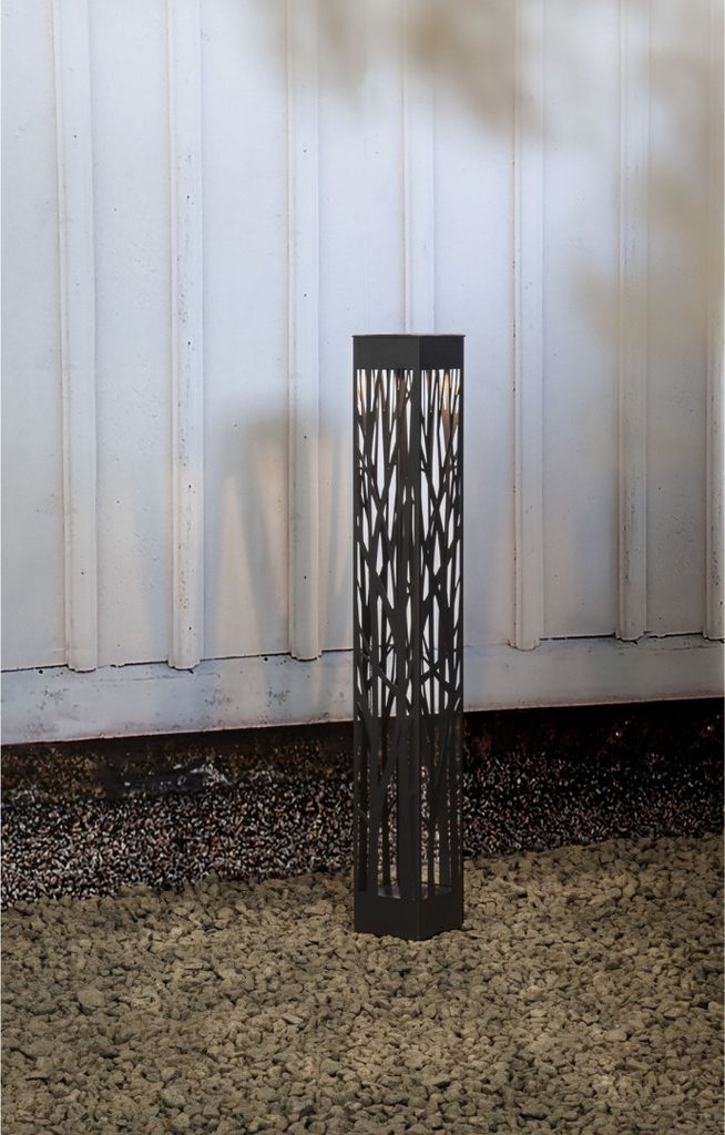 A black solar-powered LED pathway light with a decorative cut-out pattern stands on a gravel surface in front of a white corrugated wall.