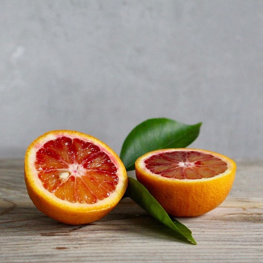 Two halves of a blood orange and two green leaves are placed on a wooden surface against a gray background.