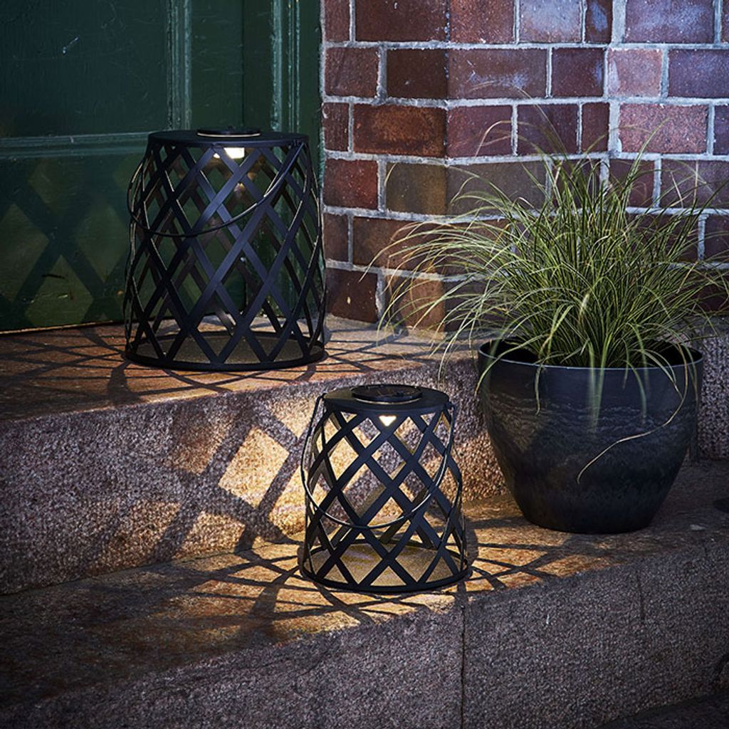 Two black metal lanterns with a diamond pattern are placed on stone steps next to a potted plant. The lanterns are lit, casting a patterned shadow on the steps.
