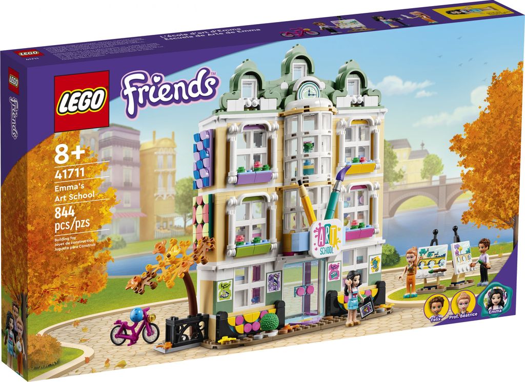 The image shows the packaging of a LEGO Friends Emma's Art School building set, displayed in a way that suggests it is a product for sale. The packaging is set against a background that includes a blurred depiction of a city scene with a river and bridge, 