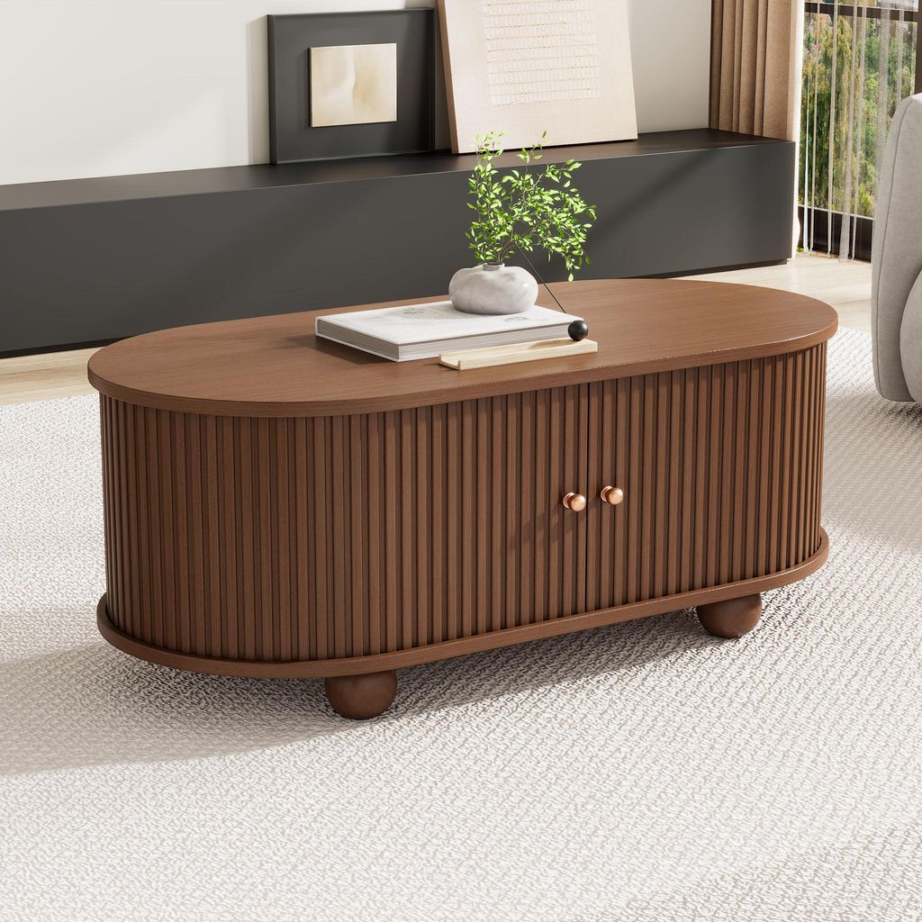 A wooden oval coffee table with ribbed doors and brass knobs sits on a textured rug in a living room setting.