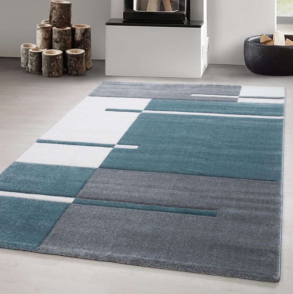 A modern geometric rug with teal, gray, and white sections is laid out on a light gray floor. In the background, a fireplace with logs and a black basket of logs are visible.