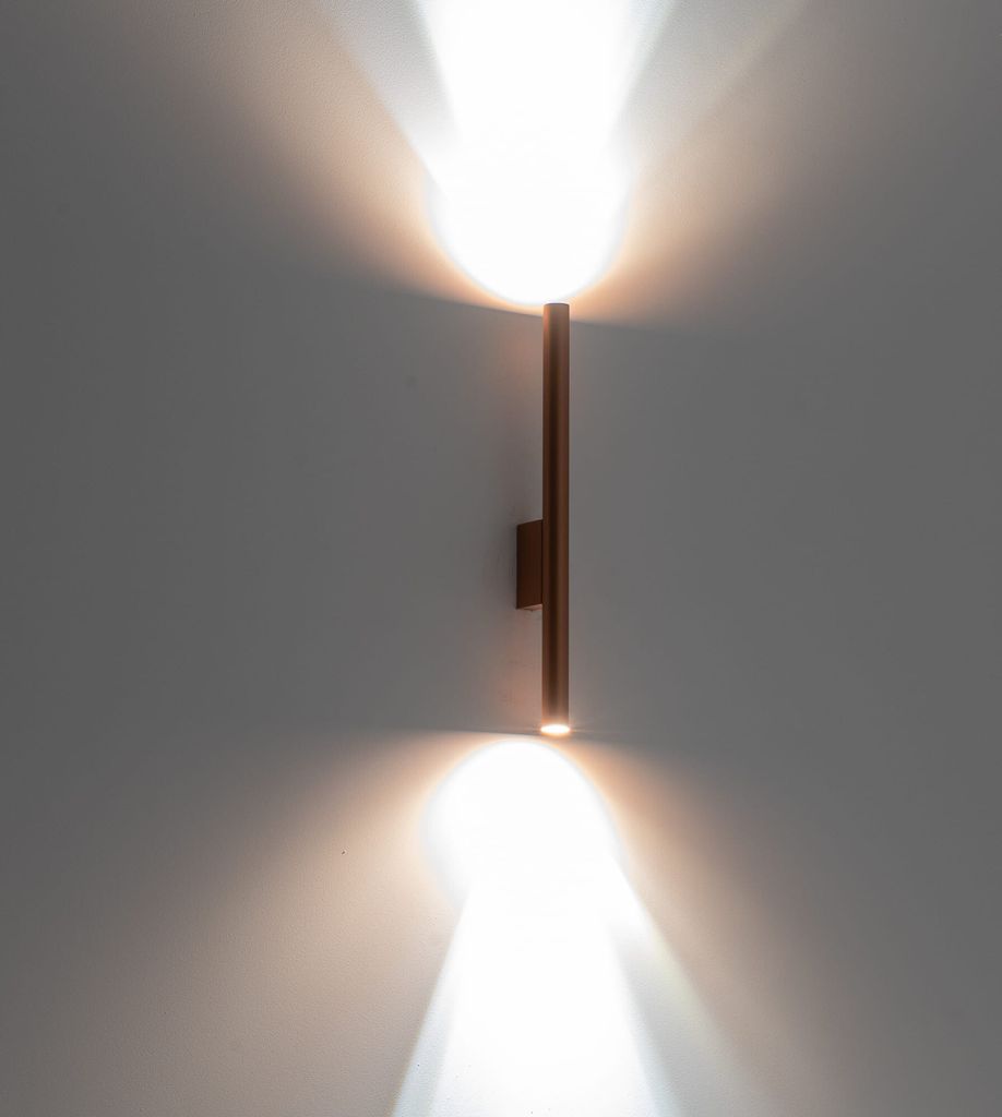 A modern copper wall sconce emitting light upwards and downwards against a gray wall.
