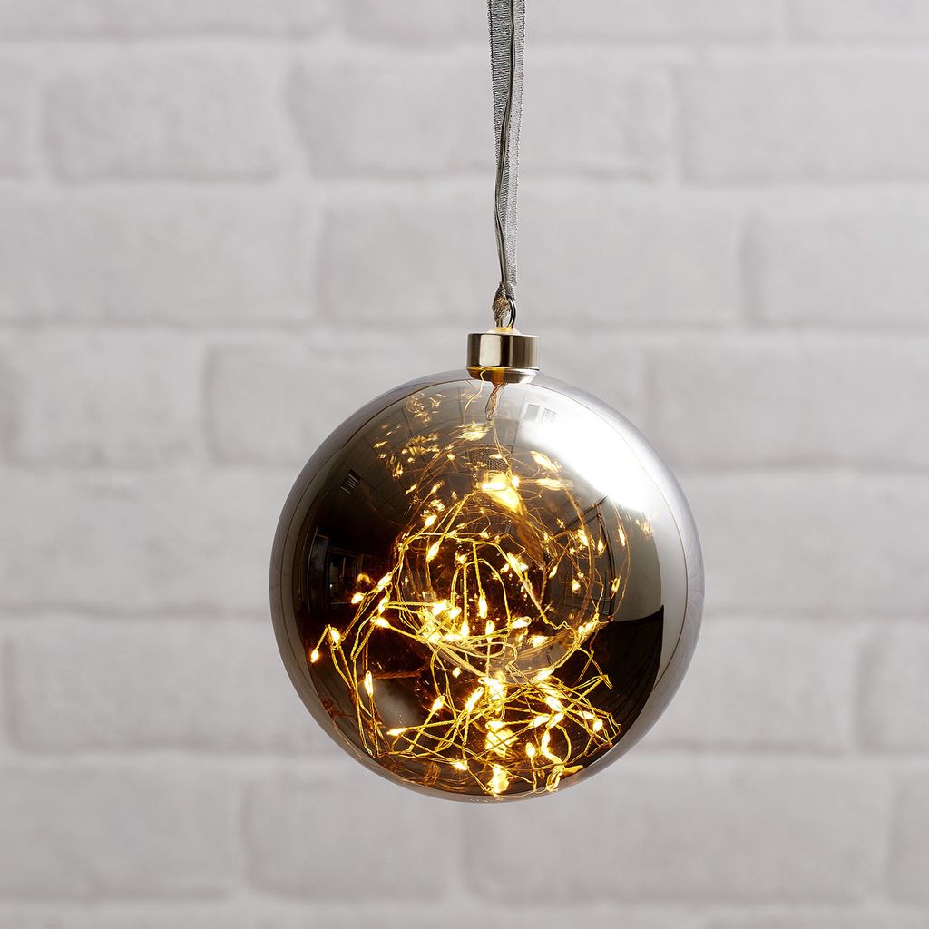 A decorative glass ornament filled with fairy lights, hanging against a white brick wall.