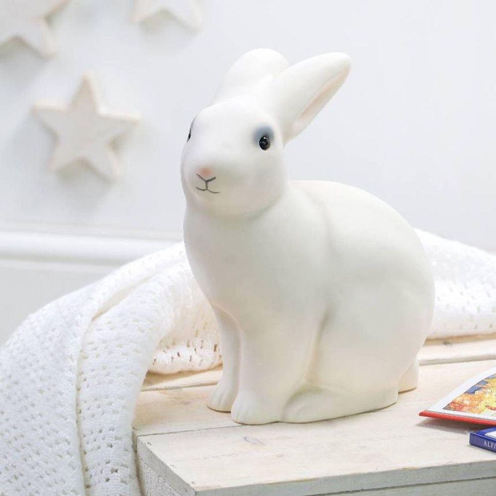A white ceramic rabbit lamp sits on a wooden crate next to a white knitted blanket and a book.