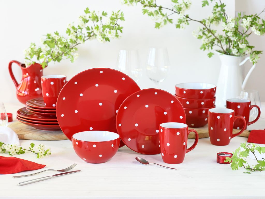 A red and white polka dot dinnerware set arranged on a table with a white pitcher and greenery.