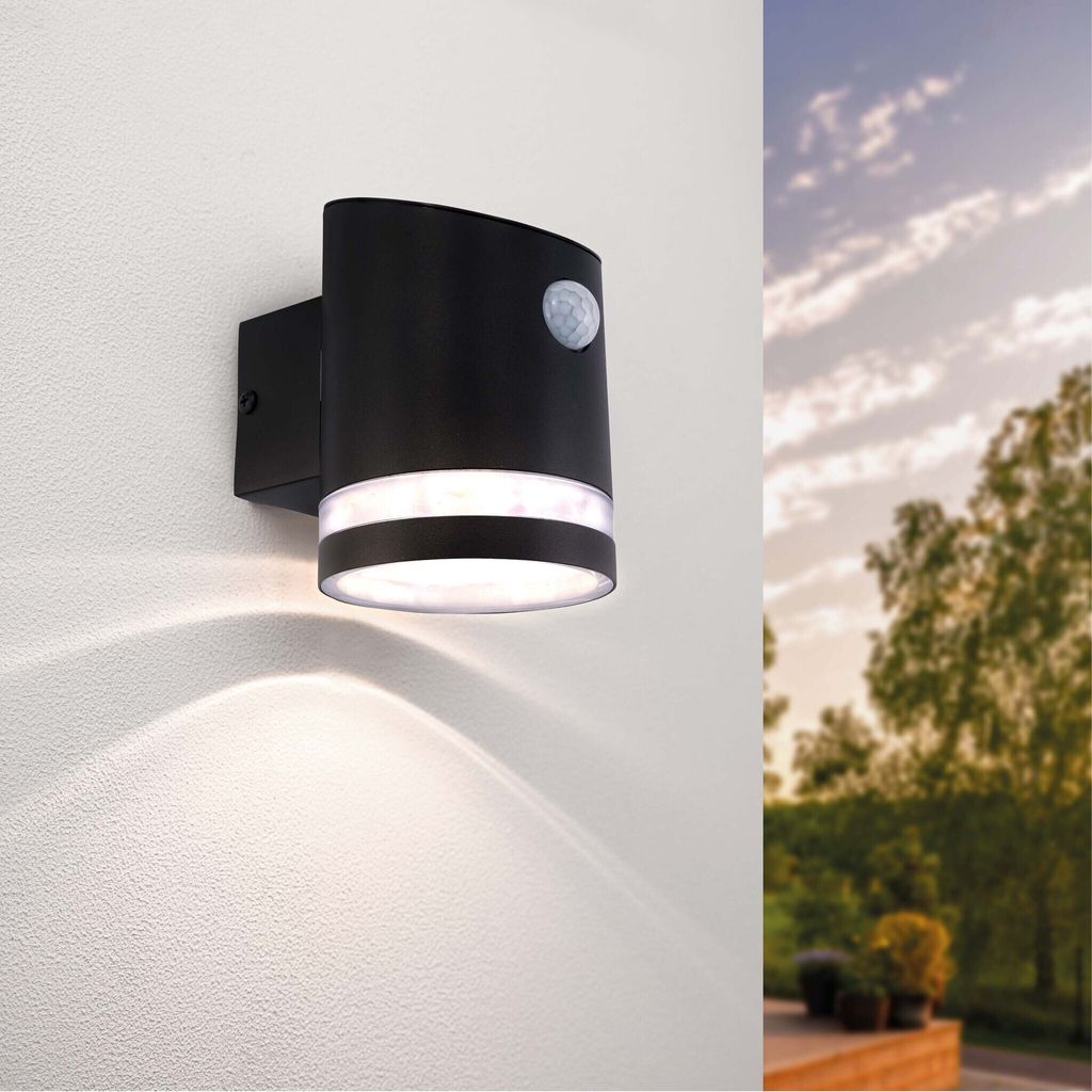 A black outdoor wall light with a motion sensor is mounted on a white wall, with trees and a cloudy sky visible in the background.