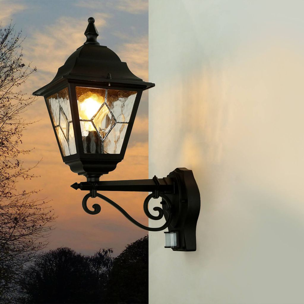 An outdoor wall lamp with a motion sensor is mounted on a white wall, with a sunset sky and trees in the background.