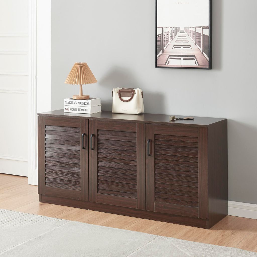 A dark brown shoe cabinet with three louvered doors is shown in a room with a light gray wall and a white door. A lamp, books, and a handbag are on top of the cabinet.
