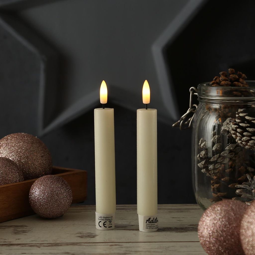 Two LED candles with a rustic look are placed on a wooden surface, surrounded by decorative Christmas ornaments and pine cones in a jar.