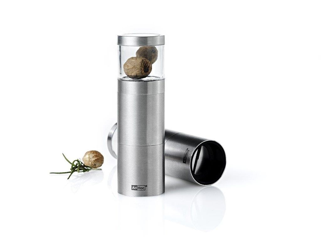 A stainless steel nutmeg grater with a glass top filled with nutmegs, placed on a white background with a sprig of rosemary and a single nutmeg to the side. Another part of the grater is lying on its side in the background.