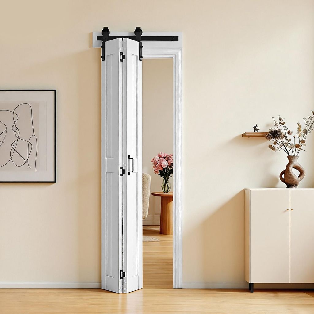 A white folding barn door is installed in a doorway, partially open, revealing a room with a vase of flowers and a side table. A framed artwork hangs on the wall to the left, and a cabinet with decorative items is on the right.