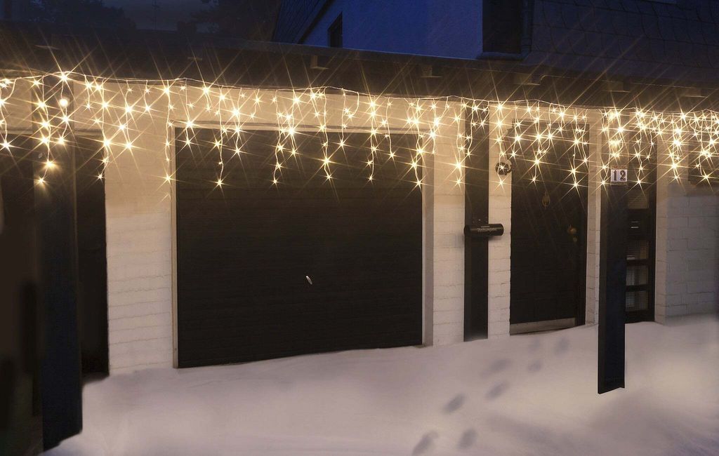 Icicle lights are hung on the eaves of a house above a garage and two doors, with snow on the ground.
