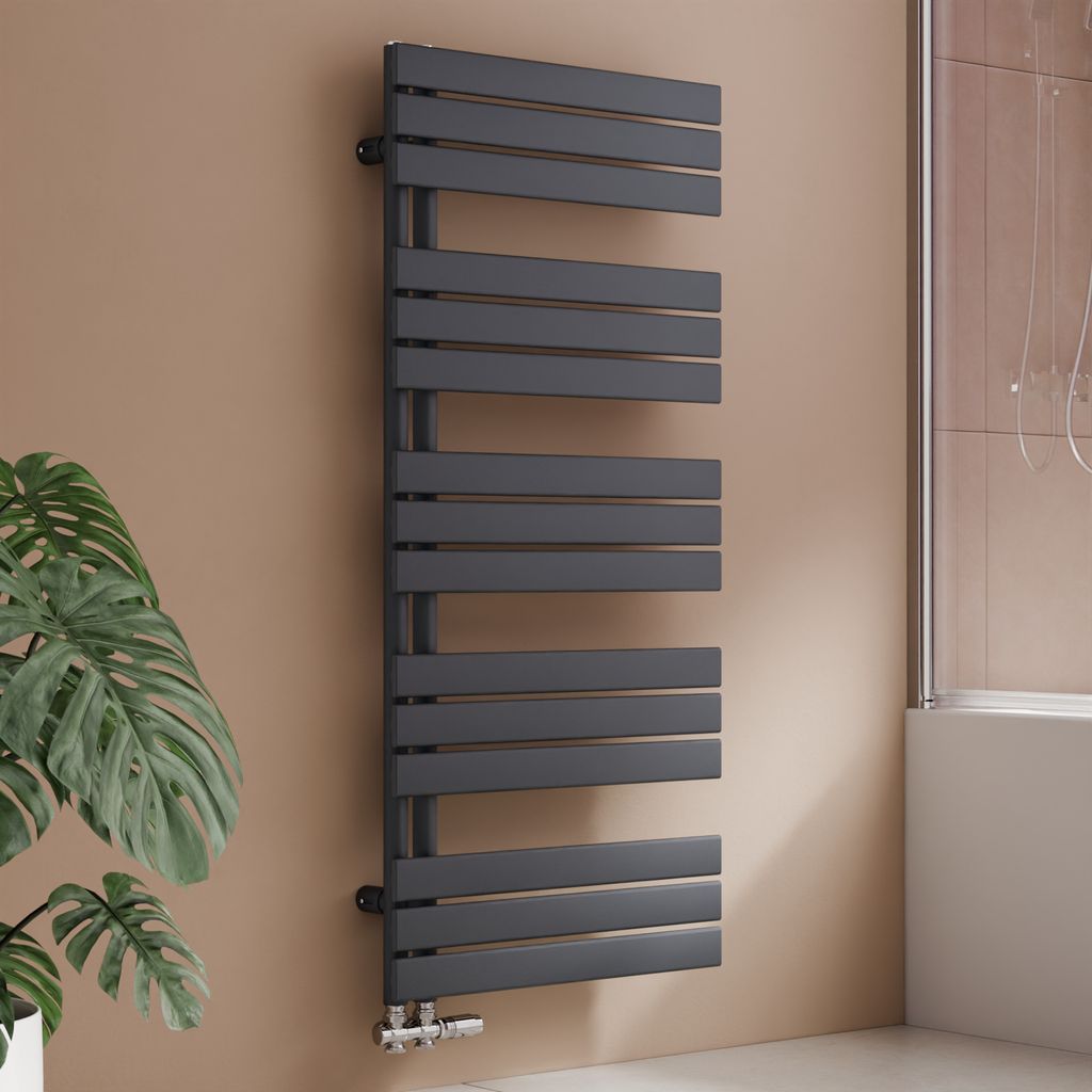 A modern anthracite radiator is mounted on a brown wall next to a large green plant and a shower enclosure.