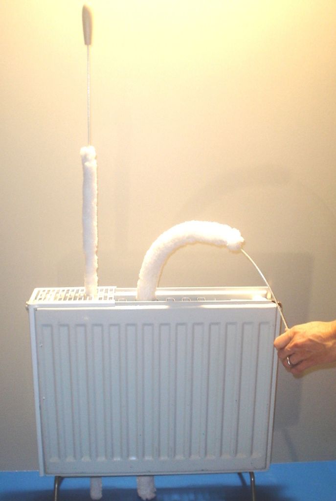 A white radiator with two cleaning tools covered in white fluffy material, one standing upright and the other curved. A hand is visible on the right side, holding a string attached to the curved tool.