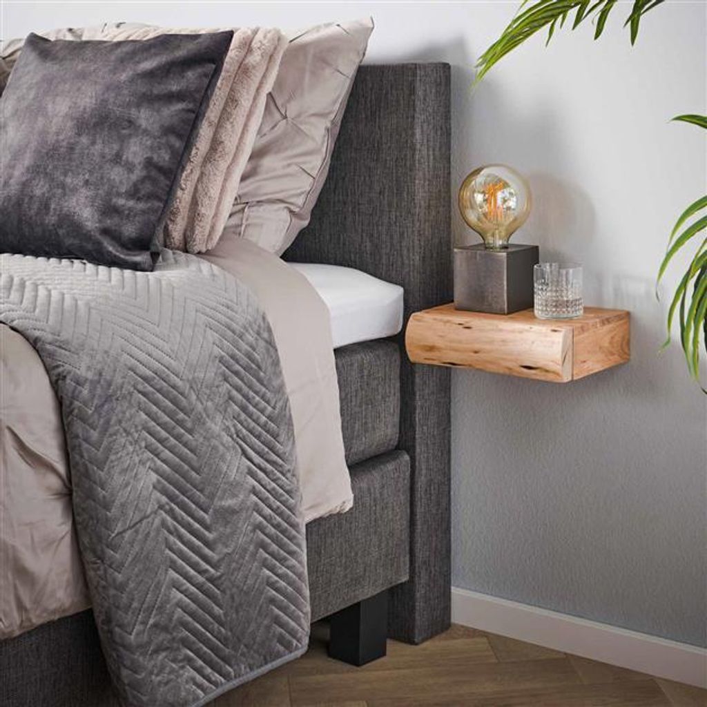 A bedside table with a lamp and a glass of water next to a bed with pillows and a blanket.