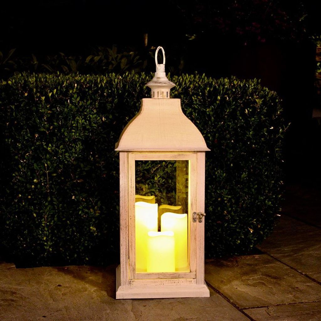 A large white lantern with three LED candles inside is placed on a stone patio in front of a dark green hedge at night.