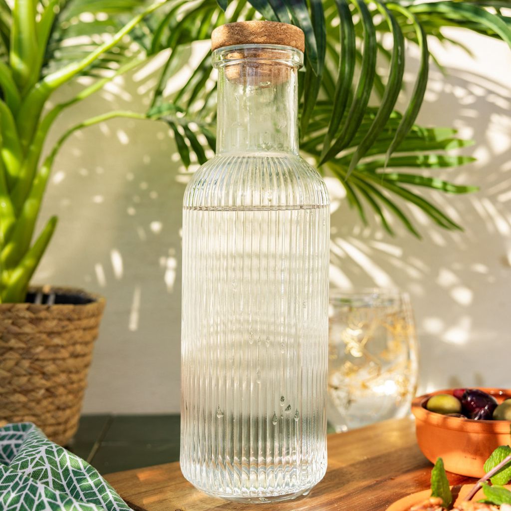 A glass carafe filled with water sits on a wooden surface, surrounded by plants and a bowl of olives.