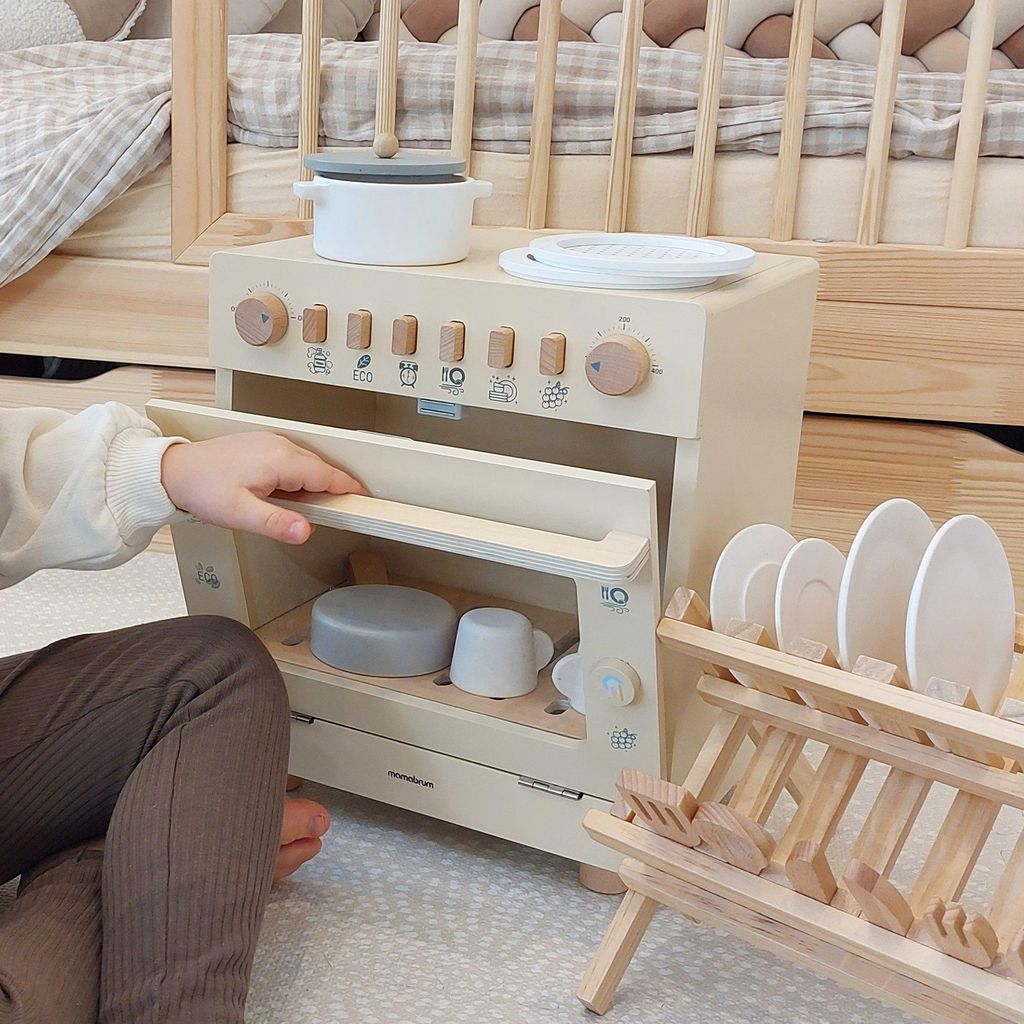 A child's hand is shown opening the door of a wooden toy dishwasher, with dishes in a drying rack nearby. The scene is set in a nursery or playroom.