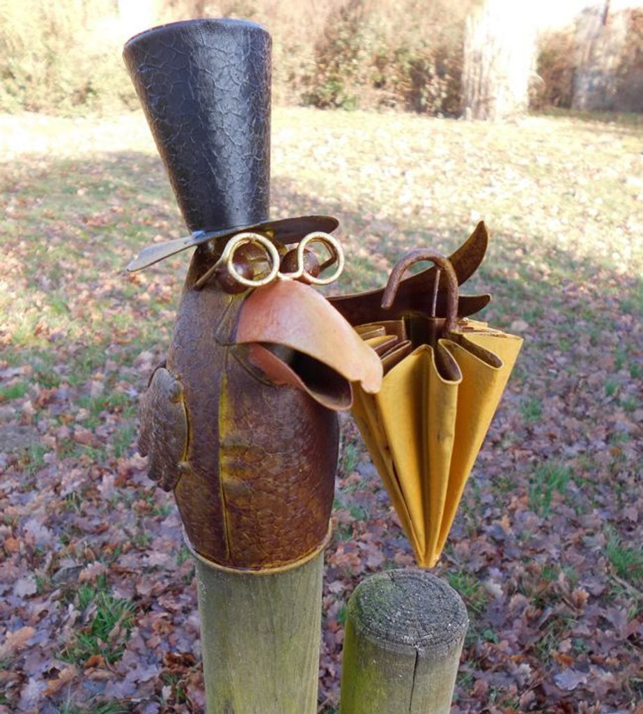 A metal sculpture of a raven wearing a top hat and glasses, holding a closed umbrella, mounted on wooden posts in an outdoor setting with fallen leaves.