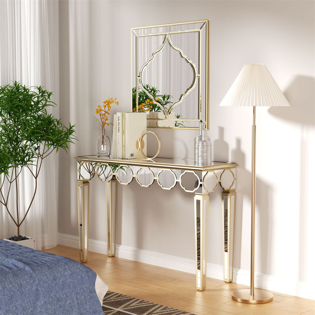 A mirrored console table with a mirror above it, decorated with books, a vase with flowers, and a decorative object, next to a floor lamp and a potted plant.