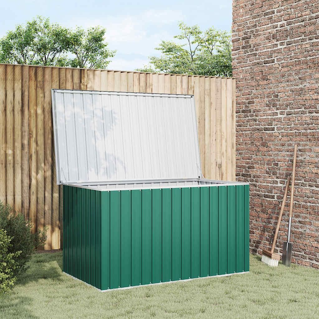 A green garden storage box with its lid open, placed on a lawn next to a wooden fence and a brick wall. Gardening tools are leaning against the wall.