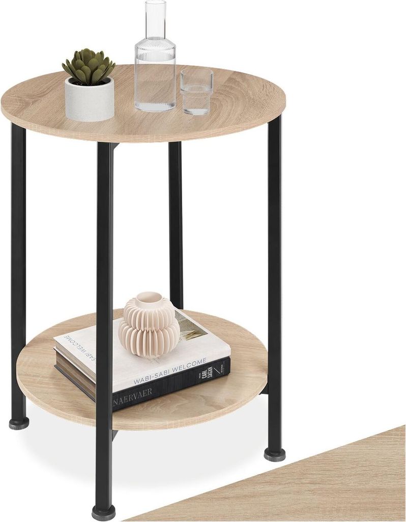 A round, two-tiered side table with a wood-look top and black metal legs, decorated with a plant, a bottle, a glass, and books.