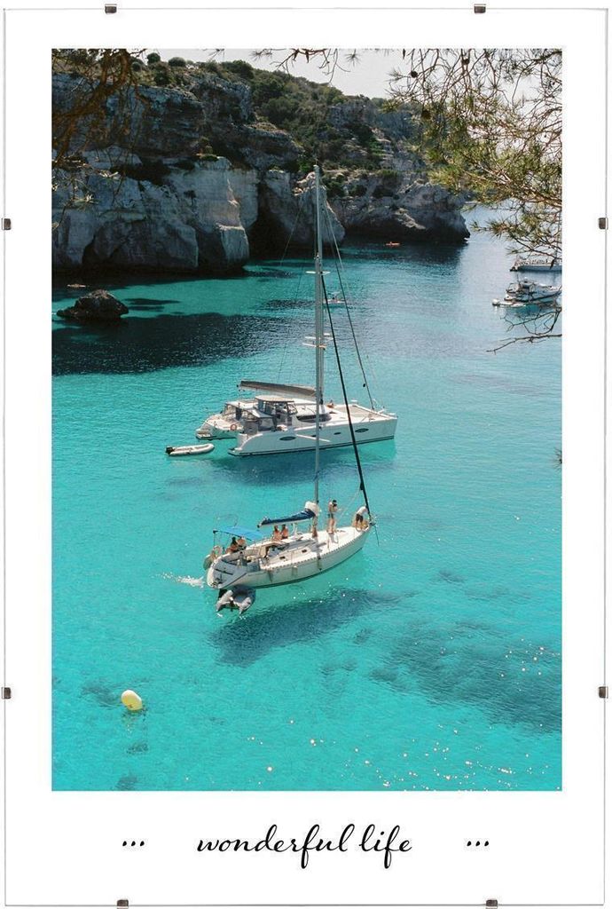 Two sailboats are anchored in a clear turquoise bay with rocky cliffs and trees in the background.