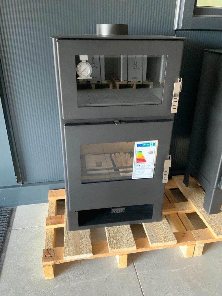 A dark gray wood-burning stove is displayed on a wooden pallet in front of a gray ribbed wall.
